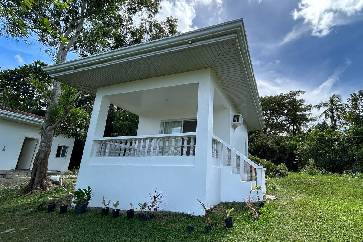 A white two-story house with a balcony, surrounded by lush greenery and trees.
