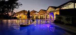 A swimming pool with a person paddleboarding, surrounded by a building with a wooden structure and glass roof. - Annabel's Resort (Ferrol)