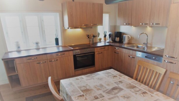 A kitchen with wooden cabinets, a dining table with a patterned tablecloth, and a window with blinds. - Holiday home Ockholm (Ockholm)