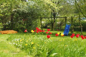 Un patio de recreo con tobogán y columpios, rodeado de césped y flores.
