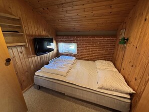 A small, wooden-paneled room with a single bed, a television mounted on the wall, and a window with curtains.