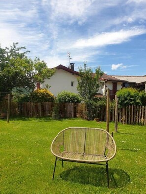 A grassy backyard with a wicker chair, a wooden fence, and a house with a tiled roof. - Gîte Elgartea arrière (St Etienne de Baigorry)