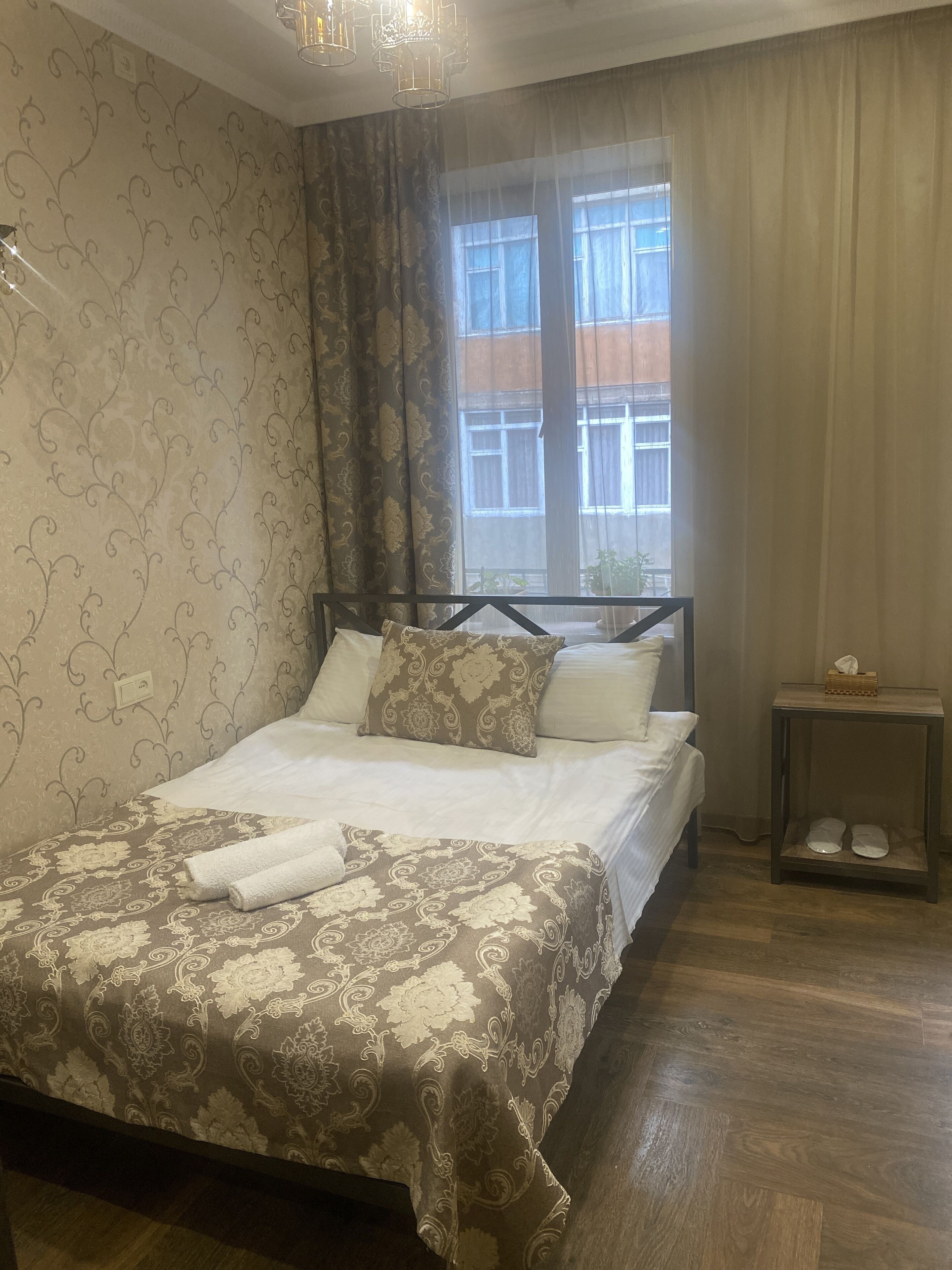 A neatly made bed with a patterned bedspread, a bedside table with a tissue box, and a window with curtains.