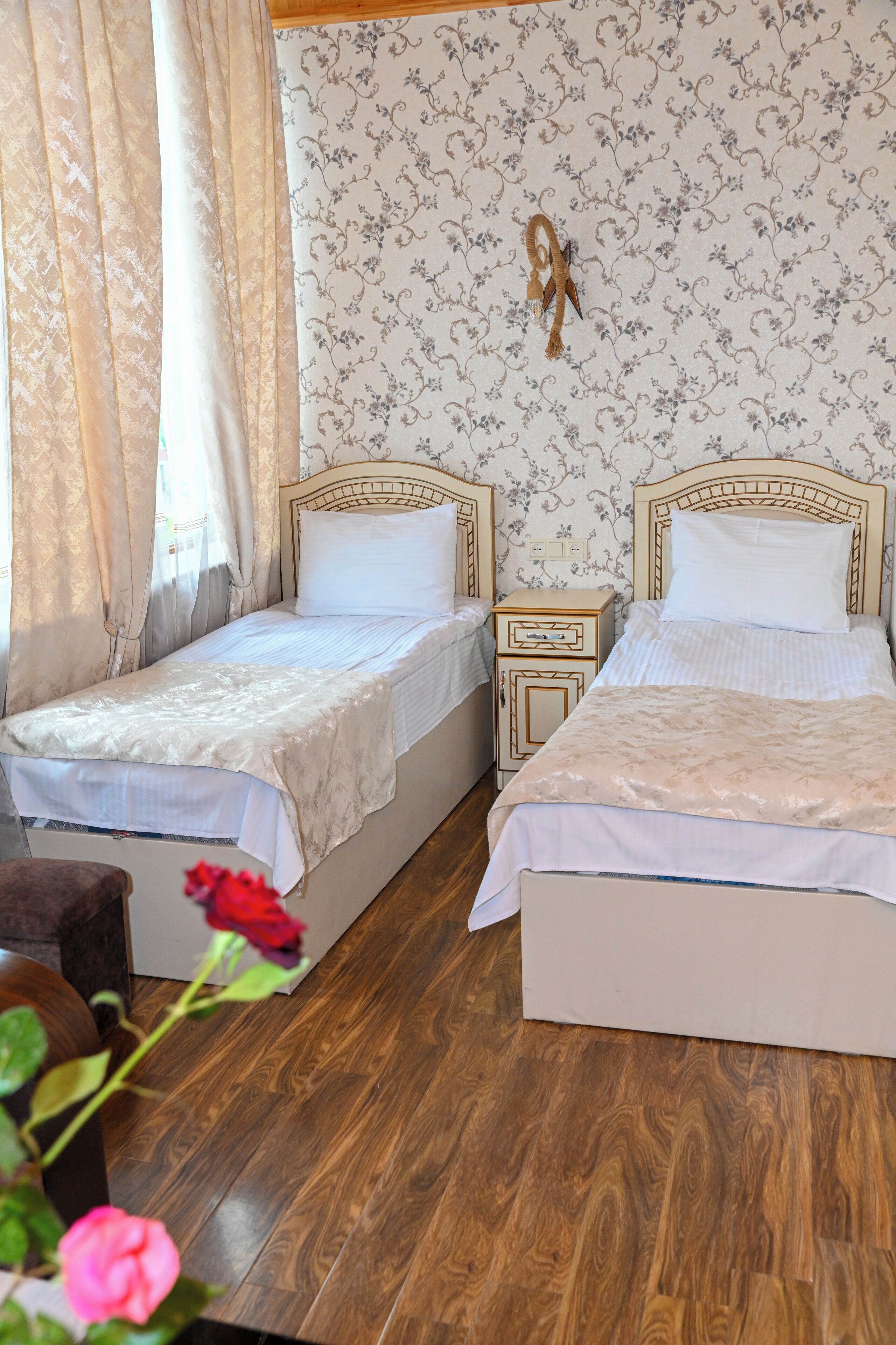 A room with two beds, wooden flooring, a floral wallpaper, and a small bedside table with a lamp.