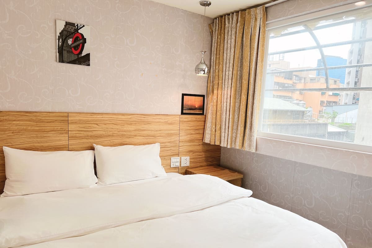 A hotel room with a wooden headboard, a bed with white bedding, a wall-mounted picture frame, a window with curtains, and a hanging pendant light.