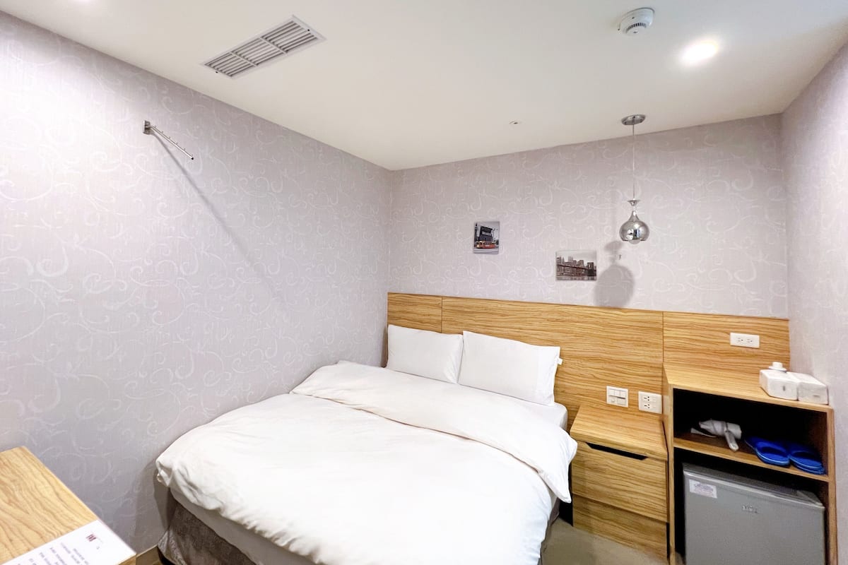 A hotel room with a bed, a nightstand, a small refrigerator, and a wall-mounted shelf with items.