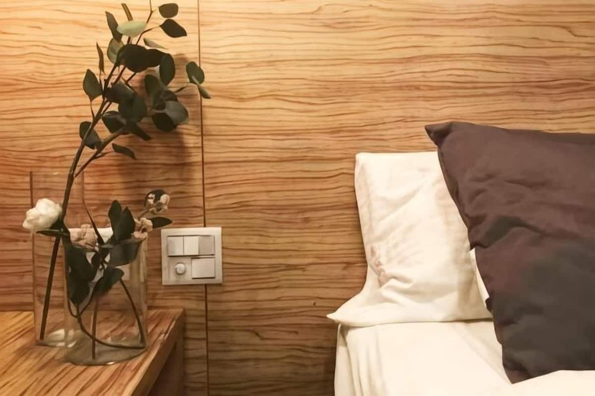 A bed with white and brown pillows, a bedside table with a glass vase containing a plant, and a wall switch.