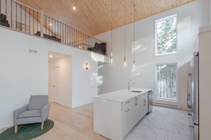 Une cuisine moderne avec un îlot central, un coin repas avec une chaise, un escalier et de grandes fenêtres.
