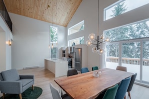 Un vaste espace de vie doté d’un plafond en bois, de grandes fenêtres, d’une table à manger et d’une cuisine située à l’arrière-plan.