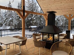 Un espace extérieur abrité, doté d’un poêle à bois, de chaises en osier et offrant une vue sur des arbres enneigés.