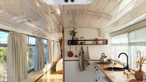 A compact kitchen with a central island, a hanging plant, and a window with curtains. - The Bus Stop (Marion Bay)