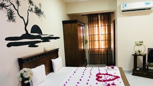 Una habitación de hotel con cama, una mesita de noche con flores, una obra de arte en la pared de un árbol y una puesta de sol, y una ventana con cortinas.
