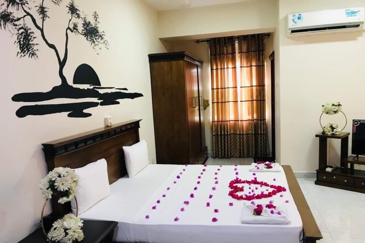 Ein Hotelzimmer mit Bett, Nachttisch mit Blumen, Wandbild mit Baum und Sonnenuntergang sowie einem Fenster mit Vorhängen.