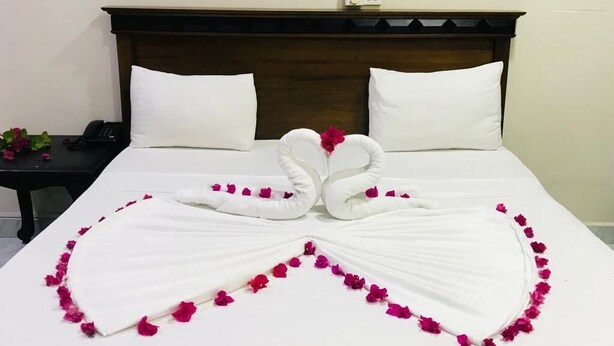 Una cama bien arreglada con una toalla dispuesta en forma de corazón y pétalos de rosa, una mesita de noche con un teléfono y una planta, y un adhesivo mural de un atardecer sobre el agua.