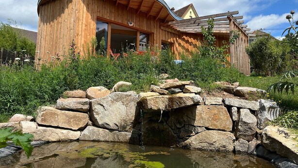 Una cabaña de madera con techo curvo, una fuente de piedra y un jardín con diversas plantas.