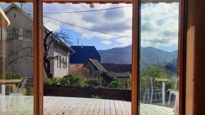 Una habitación con un amplio ventanal corredero que da a un patio exterior con terraza de madera, una mesa y vistas a una casa y montañas.