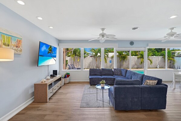 A modern living room with a sectional sofa, a flat-screen TV, a wooden cabinet, and a large window with a view of a pool and palm trees.