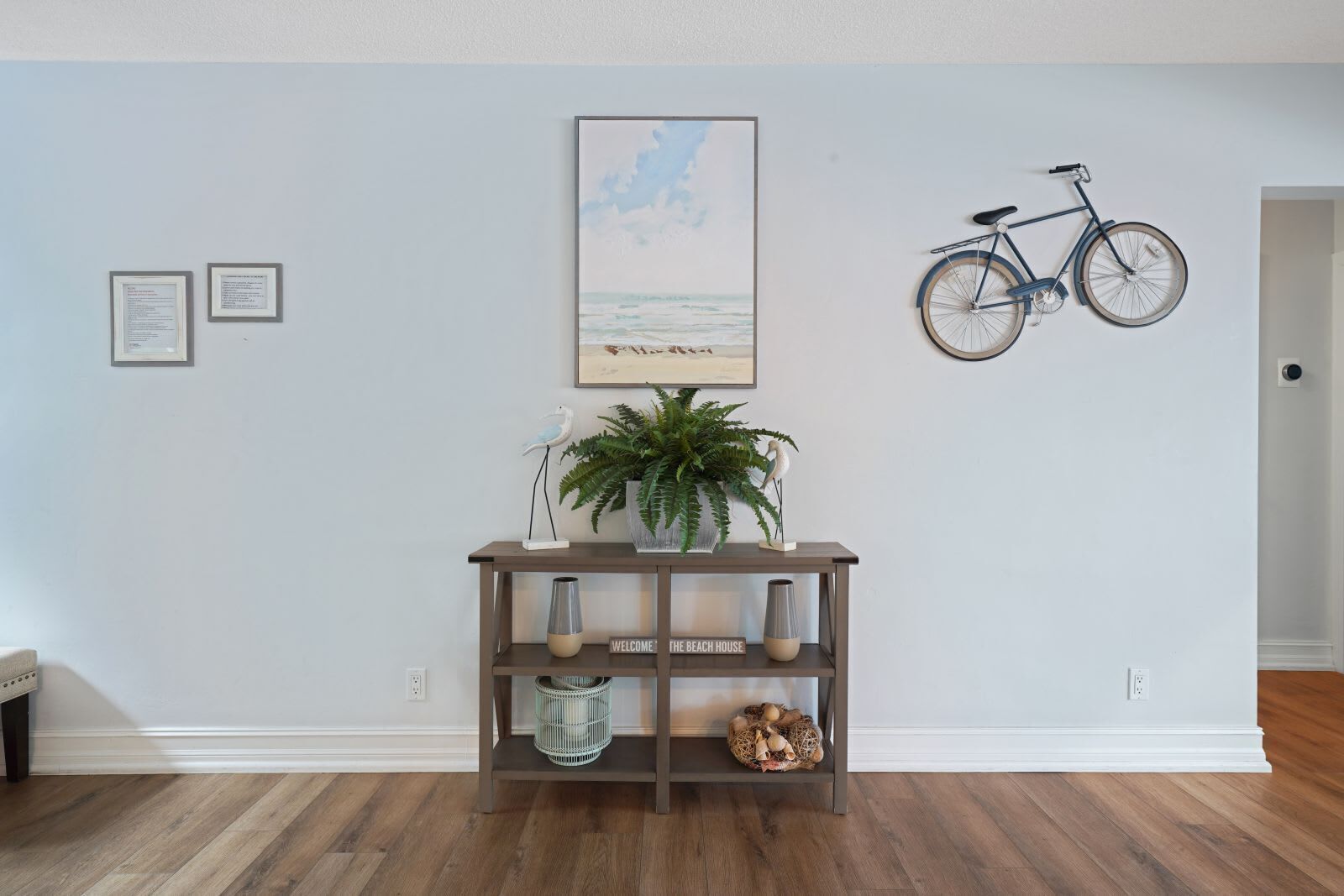 Une pièce avec un plancher en bois, une étagère fixée au mur ornée d’objets décoratifs, un tableau encadré représentant une scène de plage et un vélo accroché au mur.