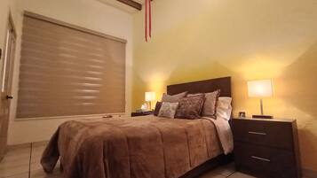 A bedroom with a bed, two bedside tables, a lamp on each table, a window with blinds, and a wooden ceiling beam.