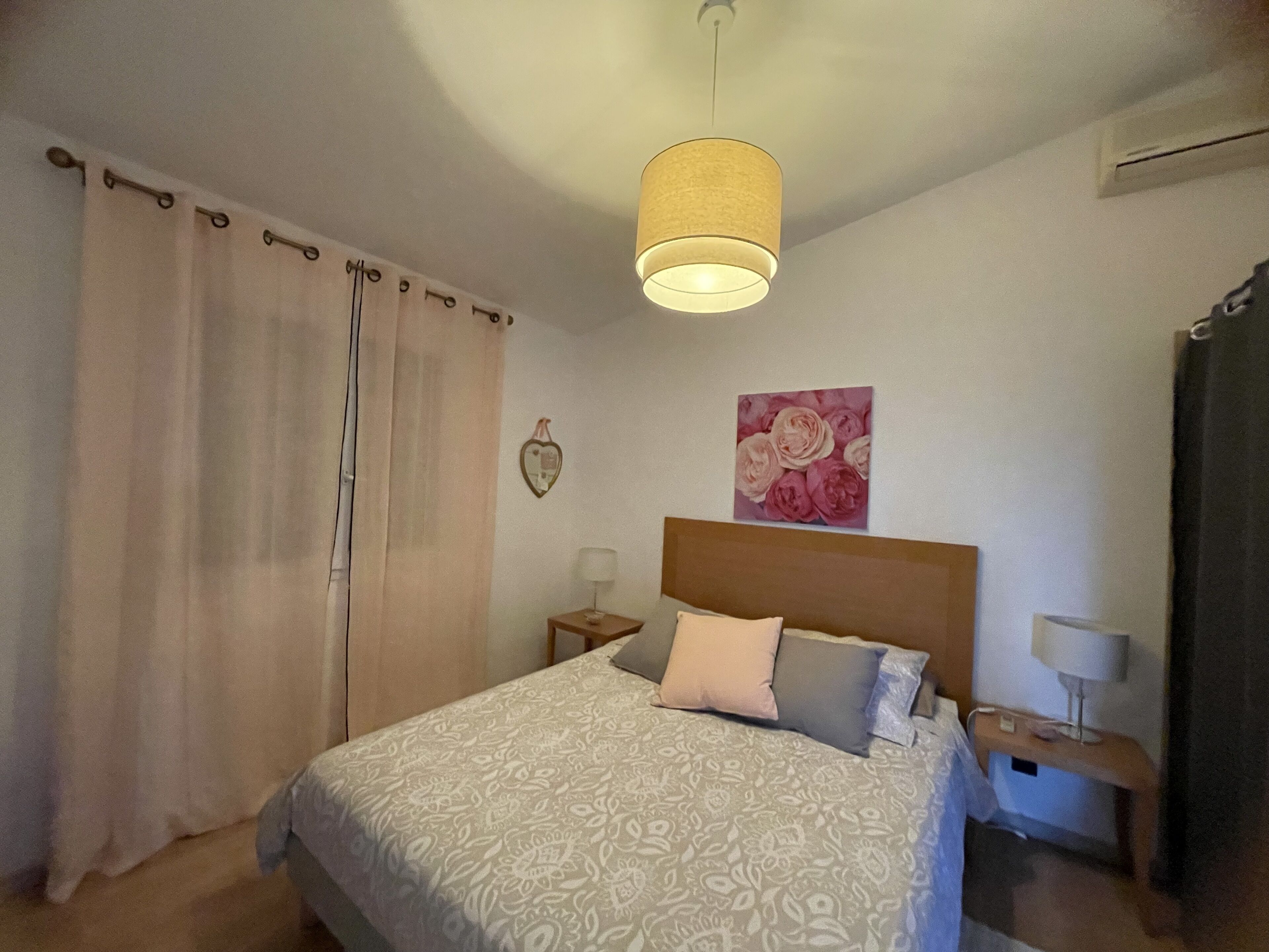 Ein Schlafzimmer mit einem Bett, einem Nachttisch, einer Lampe, einer Wanduhr und einem Fenster mit Vorhängen.