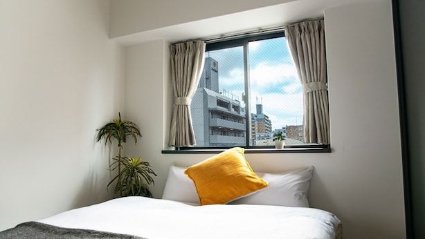 Une chambre d’hôtel avec un lit, une fenêtre avec des rideaux, une plante et une vue sur les bâtiments à l’extérieur.
