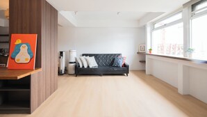 A modern living room with a black sofa, wooden flooring, and a large window with a view of buildings. - Cattail pocket Inn (Taipei)