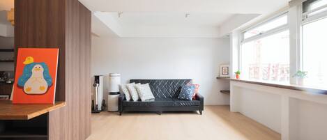A modern living room with a black sofa, wooden flooring, and a large window with a view of buildings.