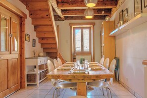 A dining area with a wooden table set for four, surrounded by chairs, in a room with wooden beams on the ceiling and a window with a view.