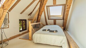 Une petite chambre à coucher de style mansardé, avec un plafond en pente, un lit simple recouvert d’une couvre-lit à motifs, un ventilateur sur pied et un puits de lumière.