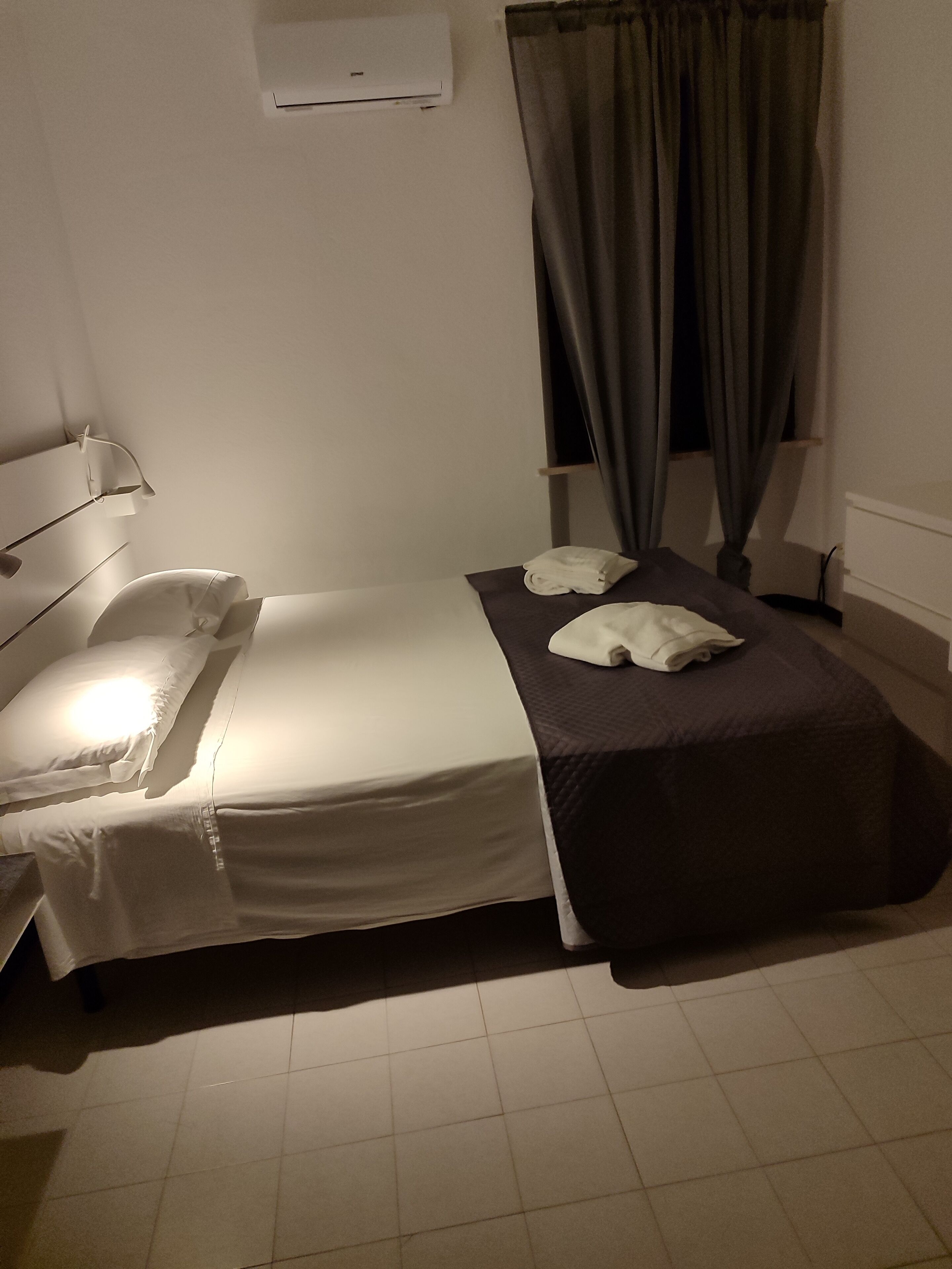 A single bed with white linens, a bedside table, a lamp, a dark-colored blanket, and a window with curtains.