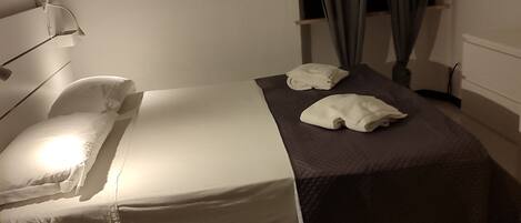 A single bed with white linens, a bedside table, a lamp, a dark-colored blanket, and a window with curtains.