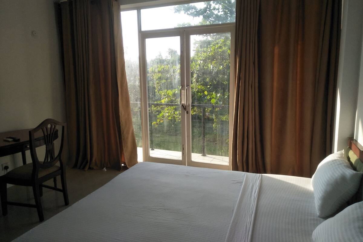 Ein Hotelzimmer mit einem großen Bett, einem Schreibtisch und einem Stuhl. Eine Glastür führt zu einem Balkon mit Blick auf Bäume.