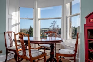A dining area with a round table and four chairs, large windows with a view of the countryside, and a red cabinet to the right.