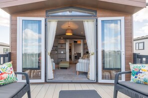 A modern outdoor living area with a sliding glass door entrance, a cushioned seating area with a bench, and a small table. - Lodge No 7 | St Andrews Lodge Park (Strathkinness)