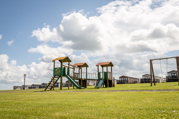 A playground with green and brown play structures, a clear sky, and houses in the background.