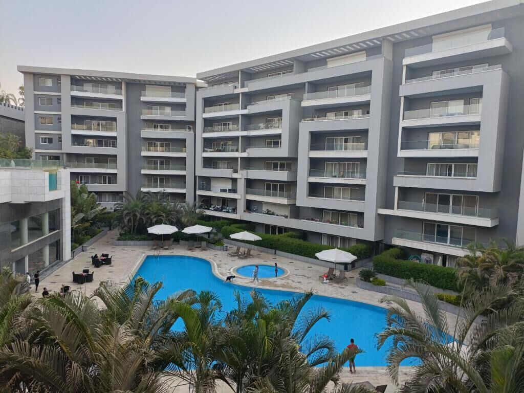 Un hotel moderno de varios pisos con una piscina central rodeada de palmeras y mobiliario exterior.