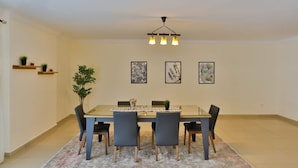 A dining area with a rectangular table, six chairs, a patterned rug, a ceiling light fixture, and three framed artworks on the wall. - Parkside boutique 3 bedroom apartment (Cairo)