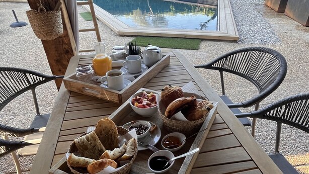 Un espacio de comedor al aire libre con una mesa de madera dispuesta para el desayuno, incluyendo pan, fruta y condimentos.