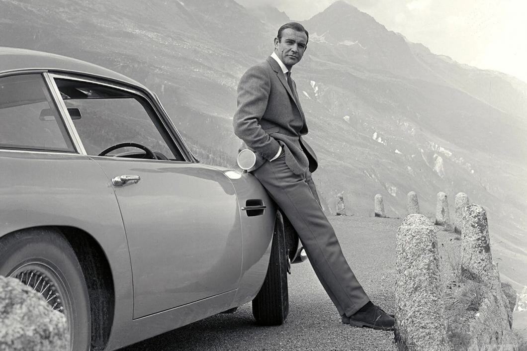 A man in a suit leaning on a vintage car with mountains in the background.