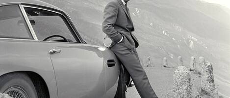 A man in a suit leaning on a vintage car with mountains in the background.