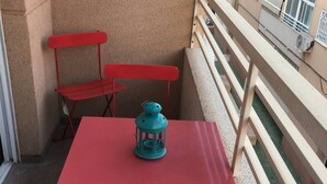 Un pequeño balcón exterior con una mesa roja y sillas a juego, un farol azul y una barandilla color beige.