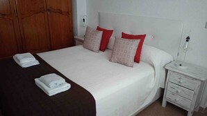 Una cama bien hecha con una colcha blanca y estampada, dos almohadas rojas y estampadas, y dos toallas blancas dobladas sobre la cama.