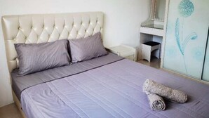 A bedroom with a tufted headboard, a bed with a grey bedspread, two pillows, a small bedside table, and a wardrobe with a floral design.
