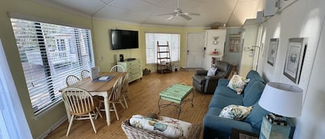 A spacious living room with a wooden floor, a dining table with chairs, a blue sofa, a television, and a ceiling fan.