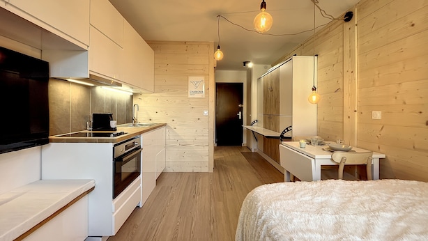 A modern kitchen with a flat-screen TV, wooden cabinets, and a dining area with a white table and chairs. - Superbe Studio Refait à Neuf en Plein Centre Ville Avec Parking (Chamonix-Mont-Blanc)