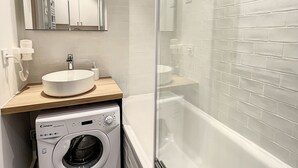 A modern bathroom with a sink, mirror, washing machine, and a glass-enclosed shower area. - Superbe Studio Refait à Neuf en Plein Centre Ville Avec Parking (Chamonix-Mont-Blanc)