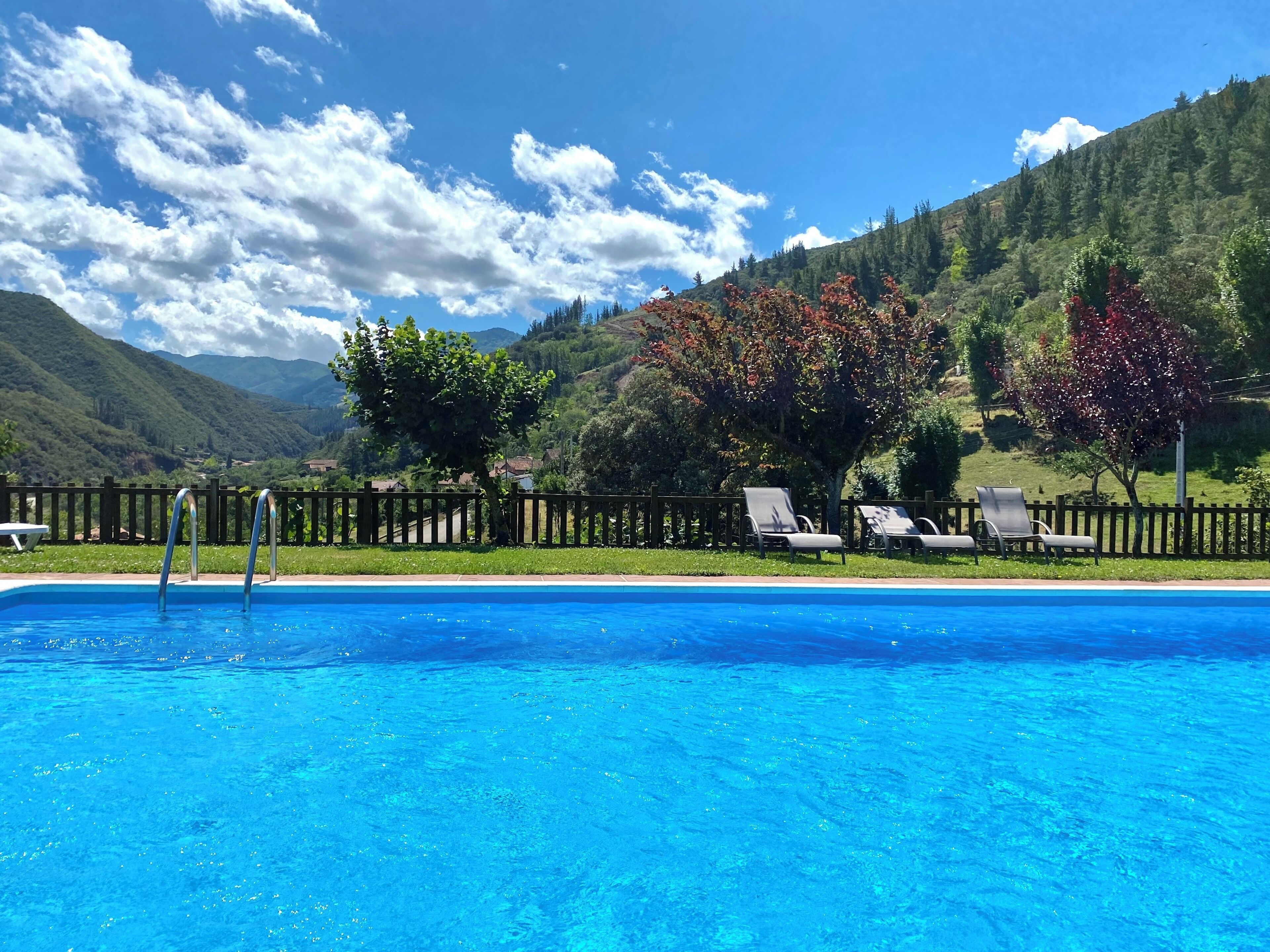 Una piscina con sillas de descanso y una valla de madera, rodeada de árboles y un paisaje montañoso.