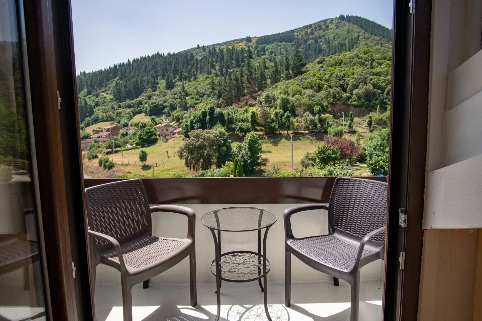 Un balcón con dos sillas de mimbre y una mesa pequeña, con vistas a un paisaje verde con colinas y árboles.
