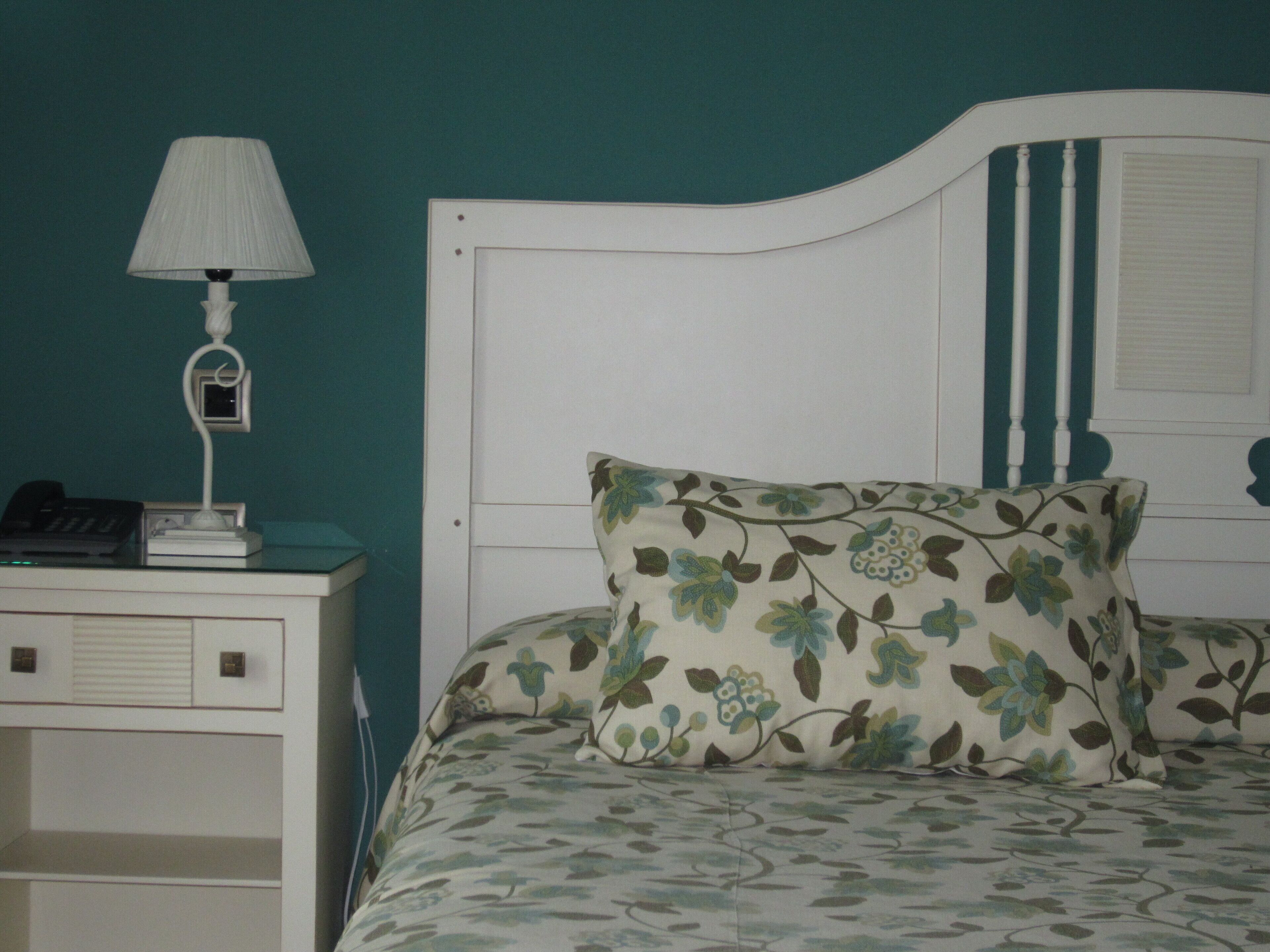 A bedroom with a white headboard, a floral-patterned pillow, a bedside table with a lamp, and a telephone.