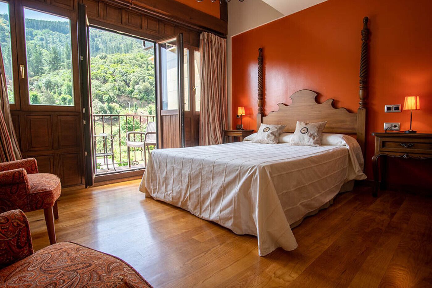 A bedroom with a large bed, wooden flooring, a balcony with a view of greenery, and a red wall.